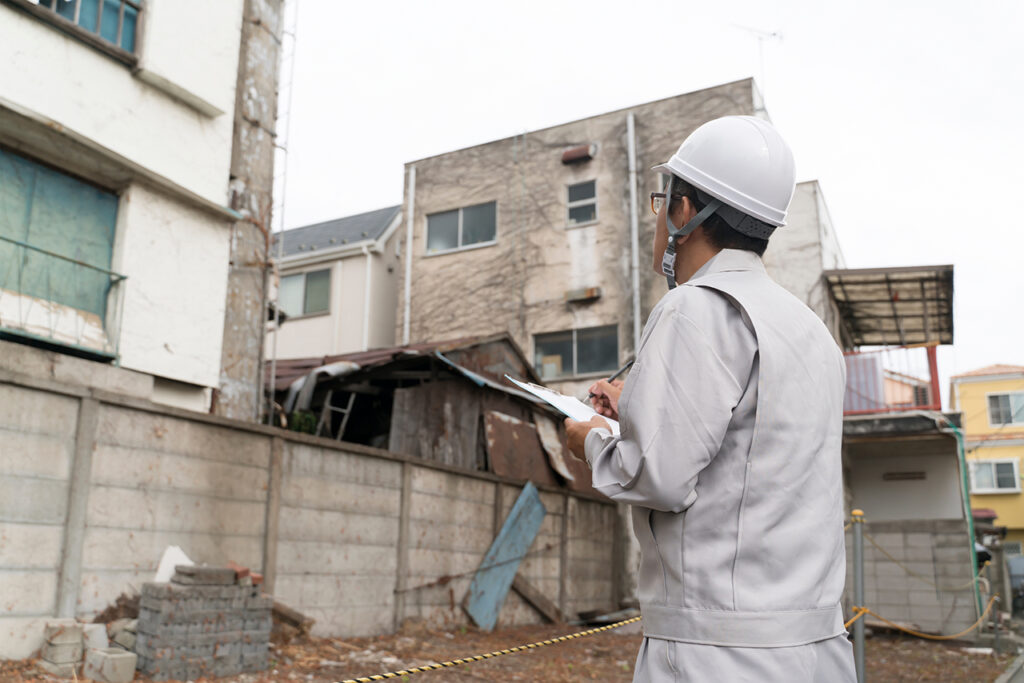 白いヘルメットと作業服姿の調査員が、工事予定の建物の前でバインダーを手に持ちながら外観を目視確認している