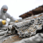 防護服、ヘルメット、全面マスクを着用した二人の人物が、下からのアングルで古びたスレート屋根の上で作業している。