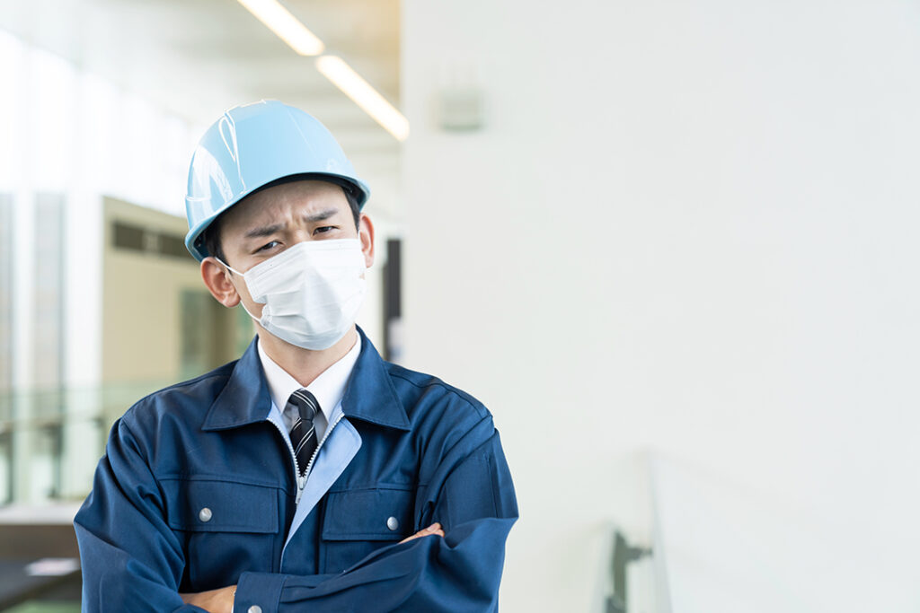 白いマスクと水色のヘルメットを着用した、作業服姿の男性。男性は腕を組み、眉間にシワを寄せ、複雑な表情で何かを考えている。