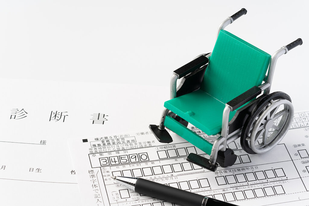 書類の上に置かれた車椅子の模型。書類のタイトルは『労働者災害補償保険』。手前には黒いボールペンが置かれている。