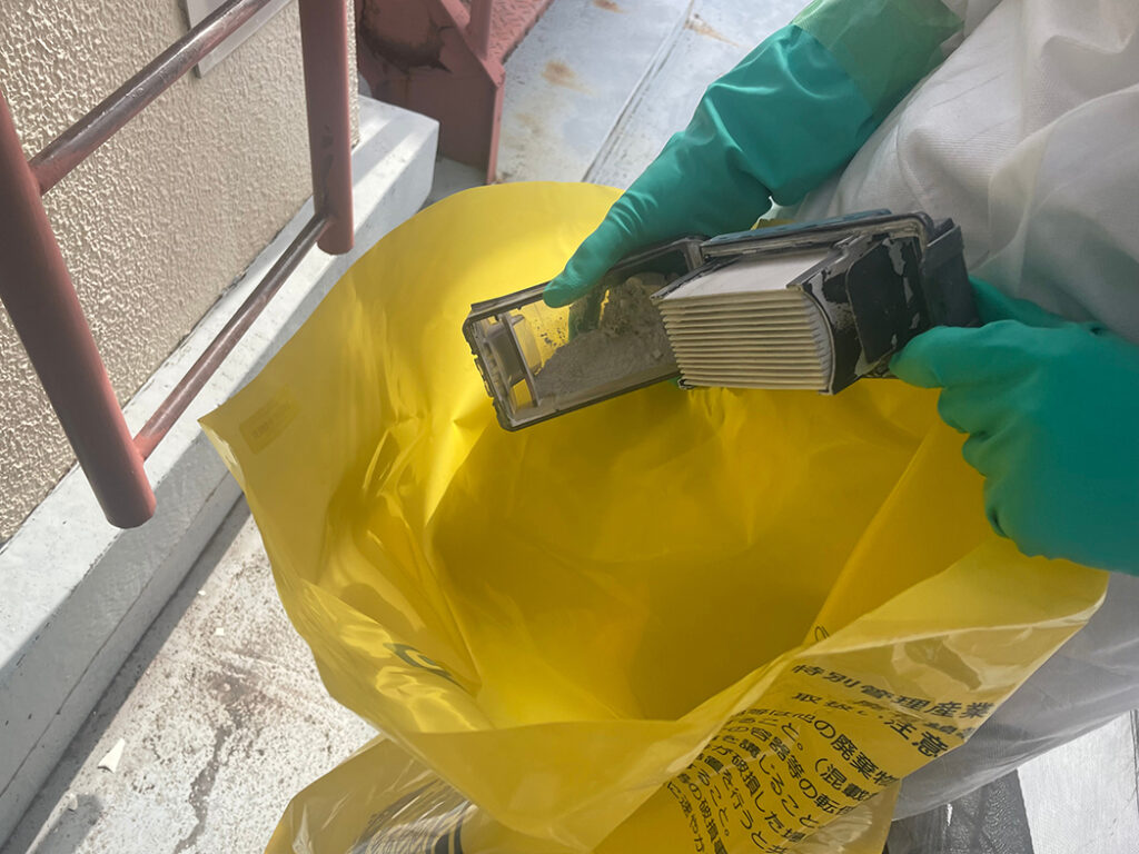 アスベスト除去現場で、防護服と緑色の手袋をした人が、除去に使用したフィルター(繊維状の粉塵が付着)を「特別管理産業廃棄物」と書かれた黄色い廃棄物袋に捨てようとしている