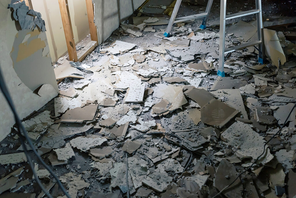 天井材が撤去され、床一面にアスベストを含有する可能性のある古い建材の破片が散乱している解体現場の室内。脚立が置かれている