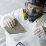 防護服とマスクを着用した作業員が、割れたレベル3アスベスト建材を専用の廃棄袋に慎重に入れている