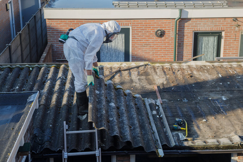 防護服とマスクを着用した作業員が、屋根の上でアスベスト含有建材である波形スレートの撤去作業を行っている